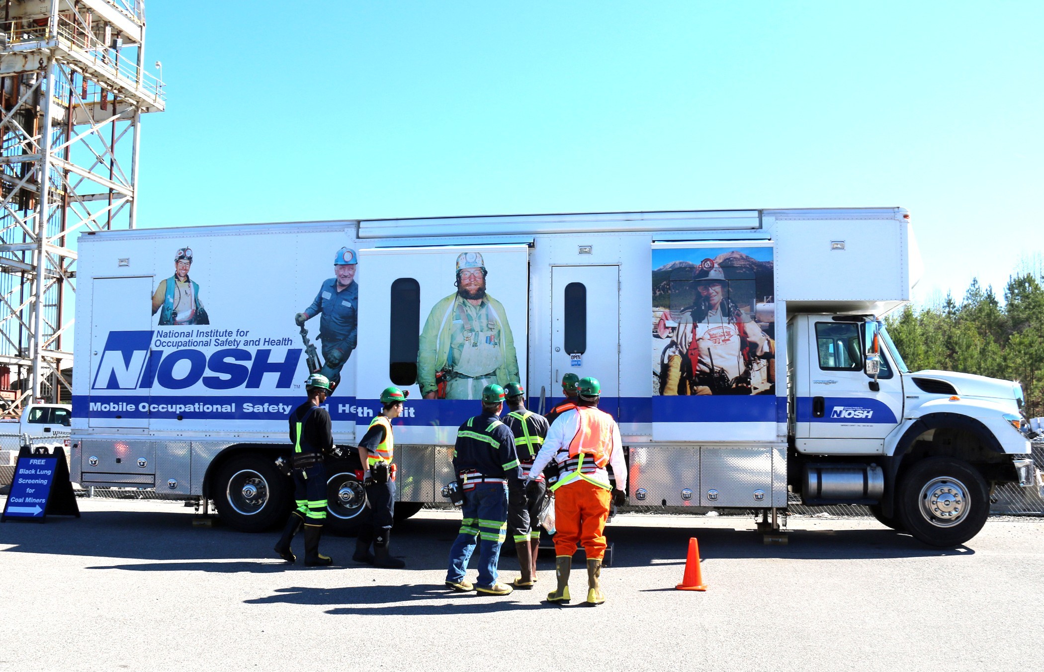 NIOSH&rsquo;s Mobile Occupational Safety and Health Unit parked outdoors, with several workers in safety gear standing in front of the vehicle. The truck displays large images of miners and the NIOSH logo. A sign nearby advertises free black lung screenings for coal miners.